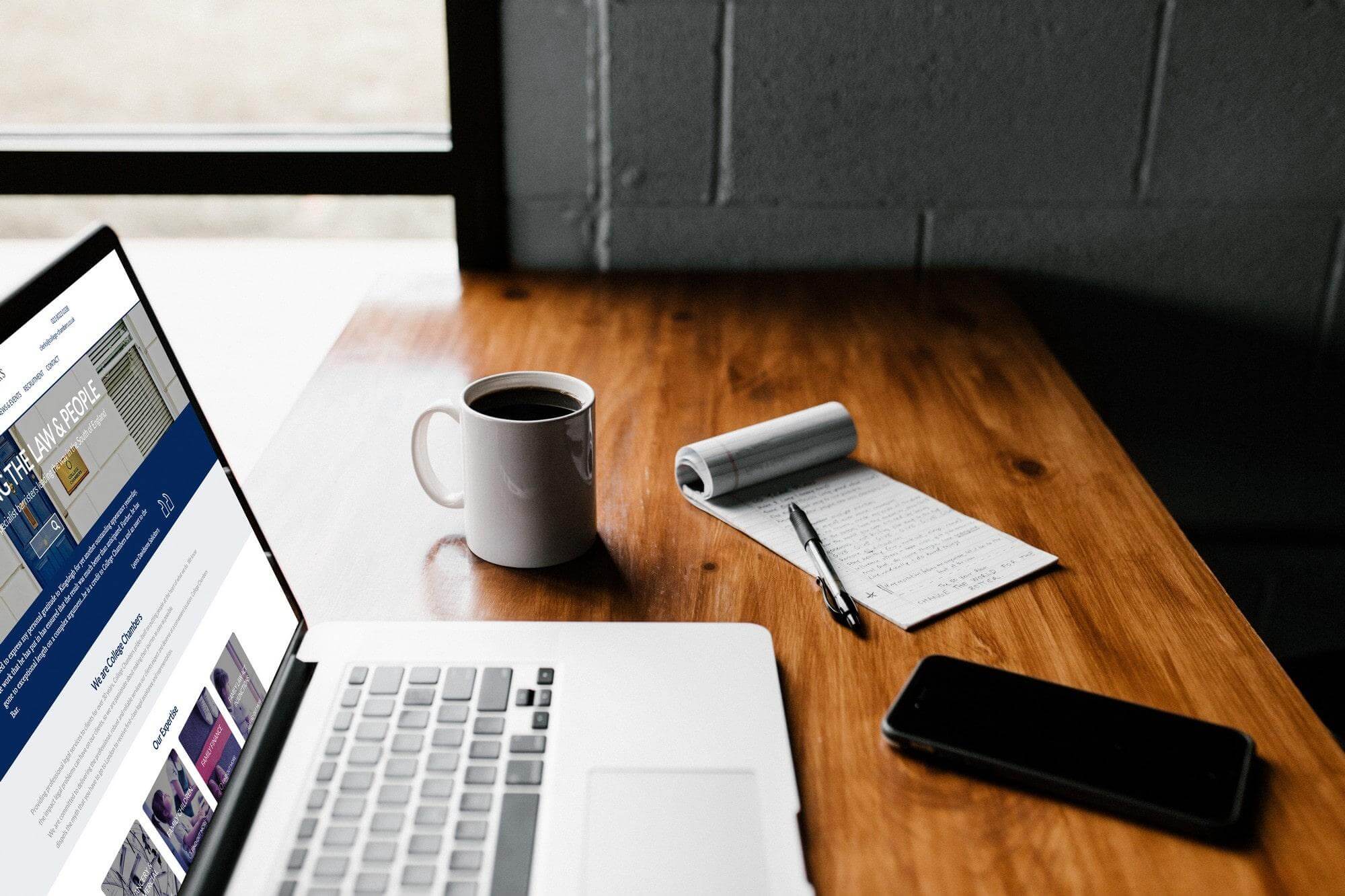 Laptop displaying website design alongside coffee mug, notepad and phone on wooden desk.