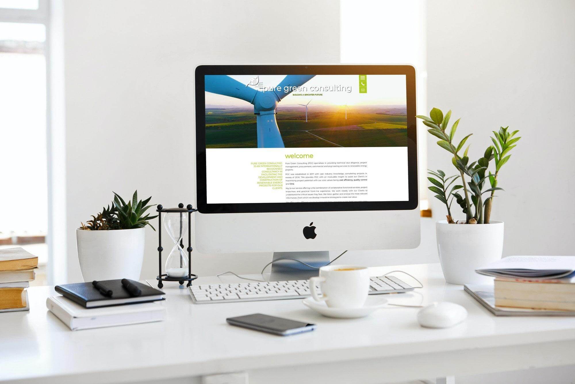 Desktop workspace with iMac displaying renewable energy website, plants, keyboard and coffee cup.