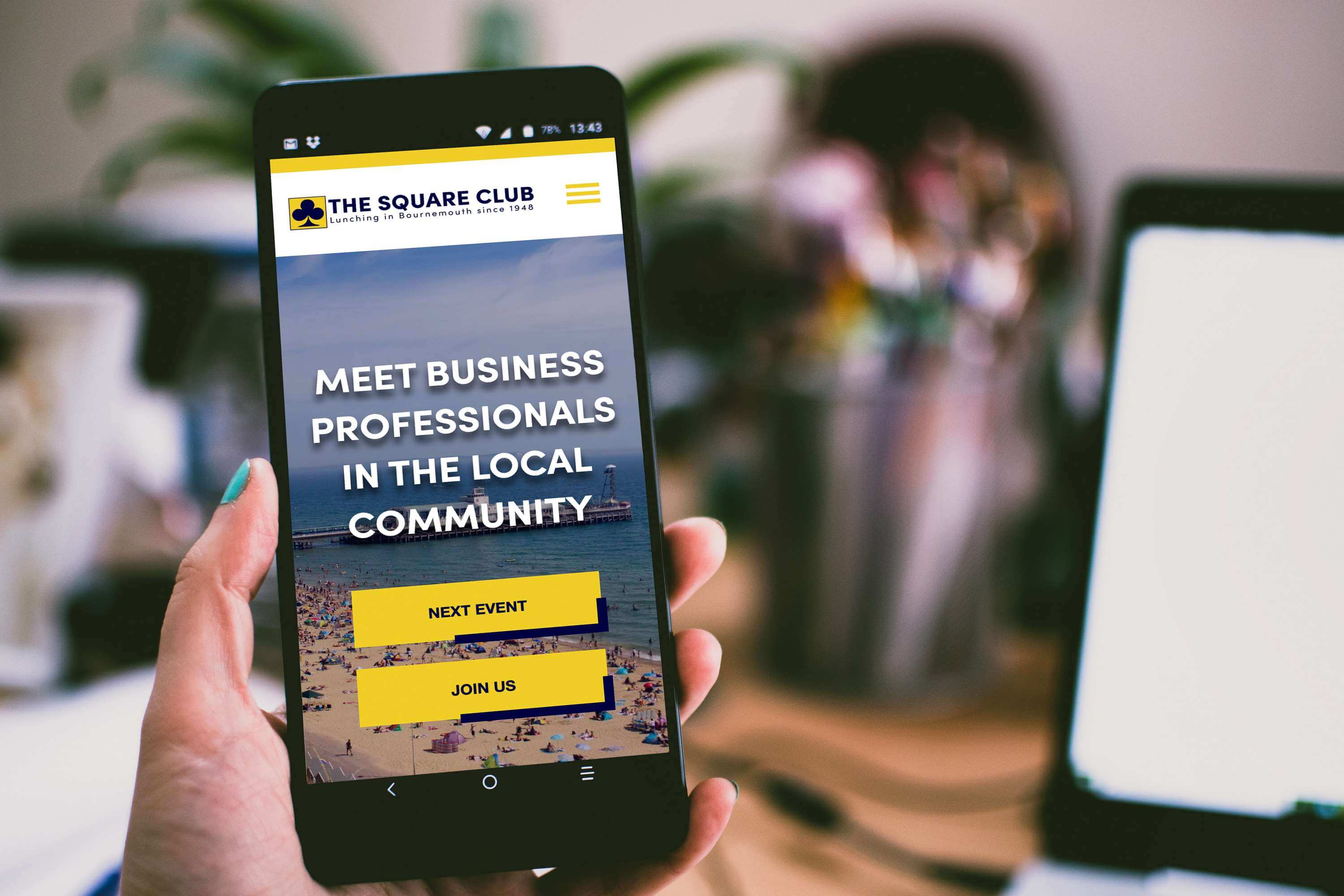 Hand holding smartphone displaying The Square Club website with networking event information and yellow call-to-action buttons.